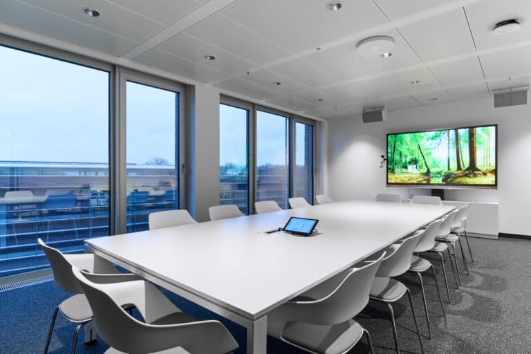 Sala de juntas ejecutiva de la Cámara de Comercio de Hannover con dos pantallas y micrófono de techo TCC M de Sennheiser, destacando la integración tecnológica para eventos corporativos híbridos.