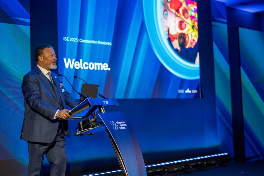 Presentación en el escenario principal de Integrated Systems Europe, con un ponente dando la bienvenida al público en un auditorio iluminado en azul.