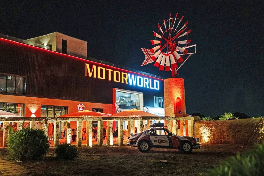 Fachada iluminada de Motorworld Mallorca por la noche, con el característico molino y zonas exteriores ambientadas mediante la instalación de audio de LD Systems.