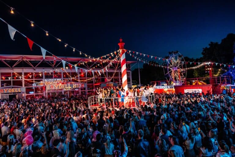 Escena de Grease de Secret Cinema en Evolution London con cientos de asistentes, escenarios iluminados y ambientación retro de Rydell High durante una experiencia inmersiva al aire libre.