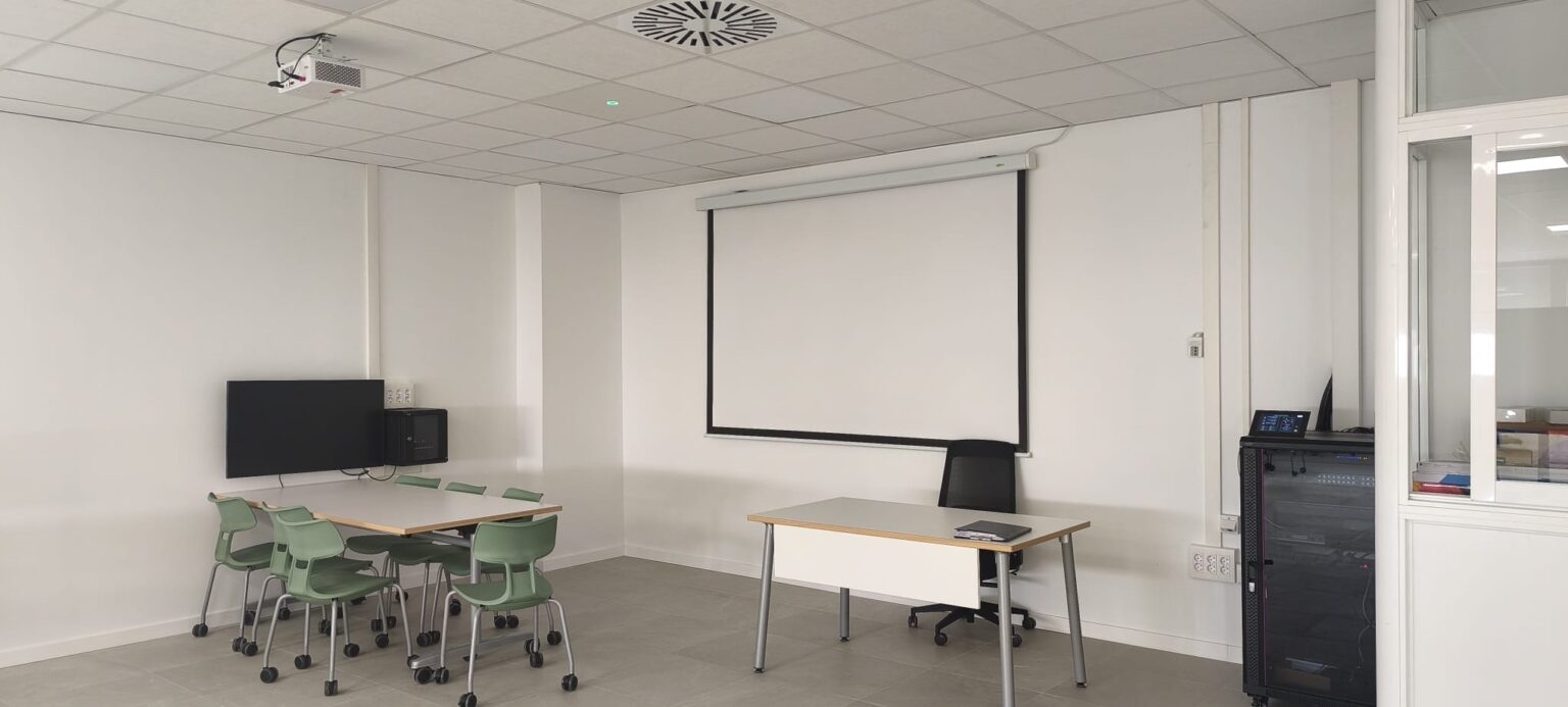 Vista general de la hiperaula Universidad de Granada equipada para docencia híbrida con tecnología audiovisual, pantalla y sistema de audio profesional.