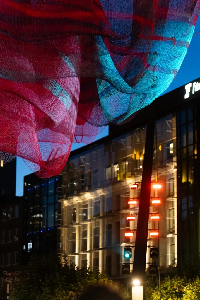 Detalle de la red de «A Sky Full of Hope» iluminada en rojo y turquesa junto a uno de los pilones que la sostienen