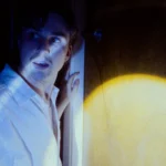 Actor apoyado en una pared de madera iluminada por una luz práctica en una escena de «Fire Alive»