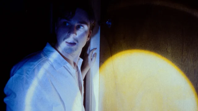Actor apoyado en una pared de madera iluminada por una luz práctica en una escena de «Fire Alive»