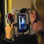Operadora de cámara sostiene un iPhone montado en un armazón mientras graba a una integrante del reparto de «Legal Queens»; en la pantalla se ve la interfaz de Blackmagic Camera
