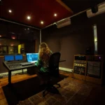 Puesto de trabajo con portátil y superficie de control en la sala docente de audio inmersivo de SAE Institute Amsterdam, con altavoces de monitorización suspendidos del techo
