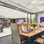 Sala de reuniones acristalada de Firmspace con mesa de juntas, butacas de piel y una pantalla corporativa mientras una persona prepara el espacio