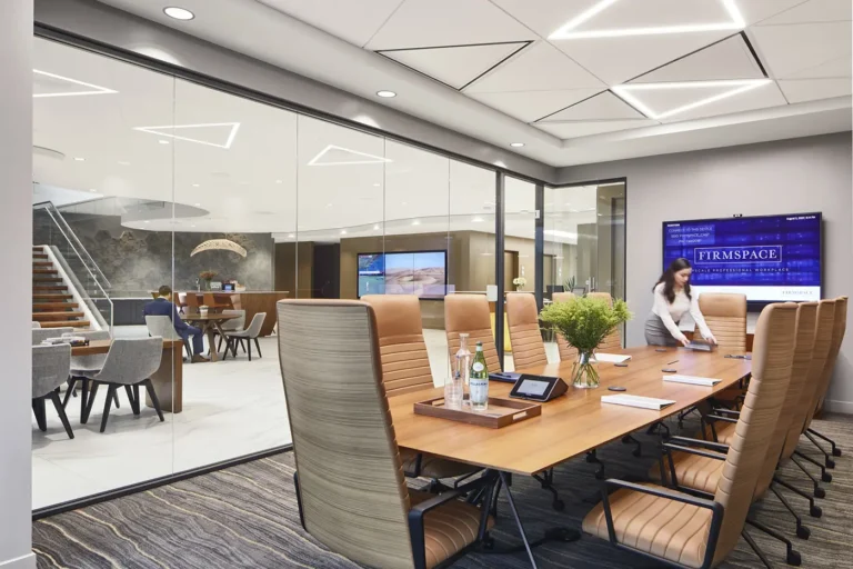 Cómo Firmspace mejora la privacidad acústica en oficinas con Biamp 5 Sala de reuniones acristalada de Firmspace con mesa de juntas, butacas de piel y una pantalla corporativa mientras una persona prepara el espacio