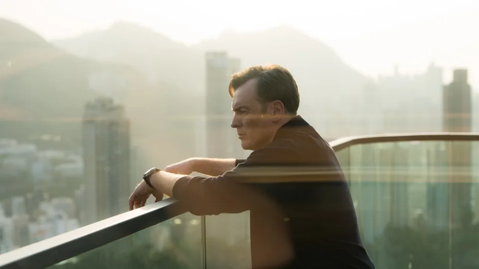 Personaje de The Season en un exterior de Hong Kong con el cielo brumoso y el perfil urbano al fondo, uno de los planos donde la coherencia del cielo condiciona el etalonaje.