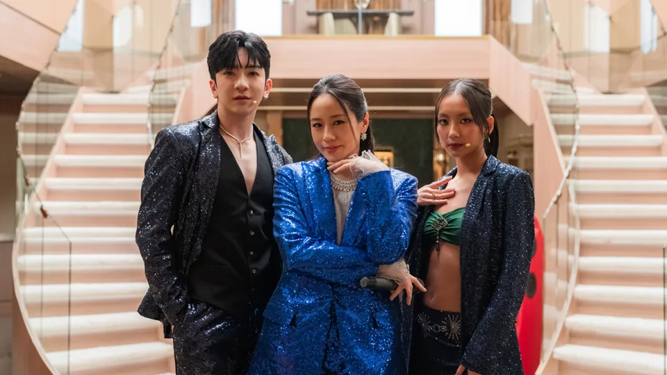 Fotograma de la serie con tres personajes de vestuario brillante en una escalinata: el etalonaje de The Season separó los tonos de piel del brillo del tejido.