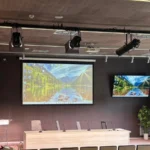 Auditorio del Hospital Universitario de Cuenca con la vara de proyectores PROLIGHTS sobre el estrado, pantalla de proyección central, dos monitores laterales, atril y mesa presidencial con micrófonos de cuello de cisne