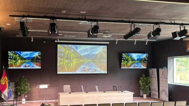 Auditorio del Hospital Universitario de Cuenca con la vara de proyectores PROLIGHTS sobre el estrado, pantalla de proyección central, dos monitores laterales, atril y mesa presidencial con micrófonos de cuello de cisne