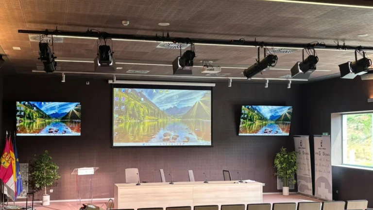 Auditorio del Hospital Universitario de Cuenca con la vara de proyectores PROLIGHTS sobre el estrado, pantalla de proyección central, dos monitores laterales, atril y mesa presidencial con micrófonos de cuello de cisne