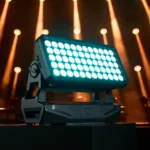 Foco wash Cameo ZENIT W1200 G2 con sus 60 LED RGBALC encendidos en tono cian sobre un pedestal, ante un escenario con haces de luz ámbar