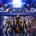 Escena coral del musical Cinderella de Lion Musicals con el elenco bajo una gran estructura de hierro iluminada en azul